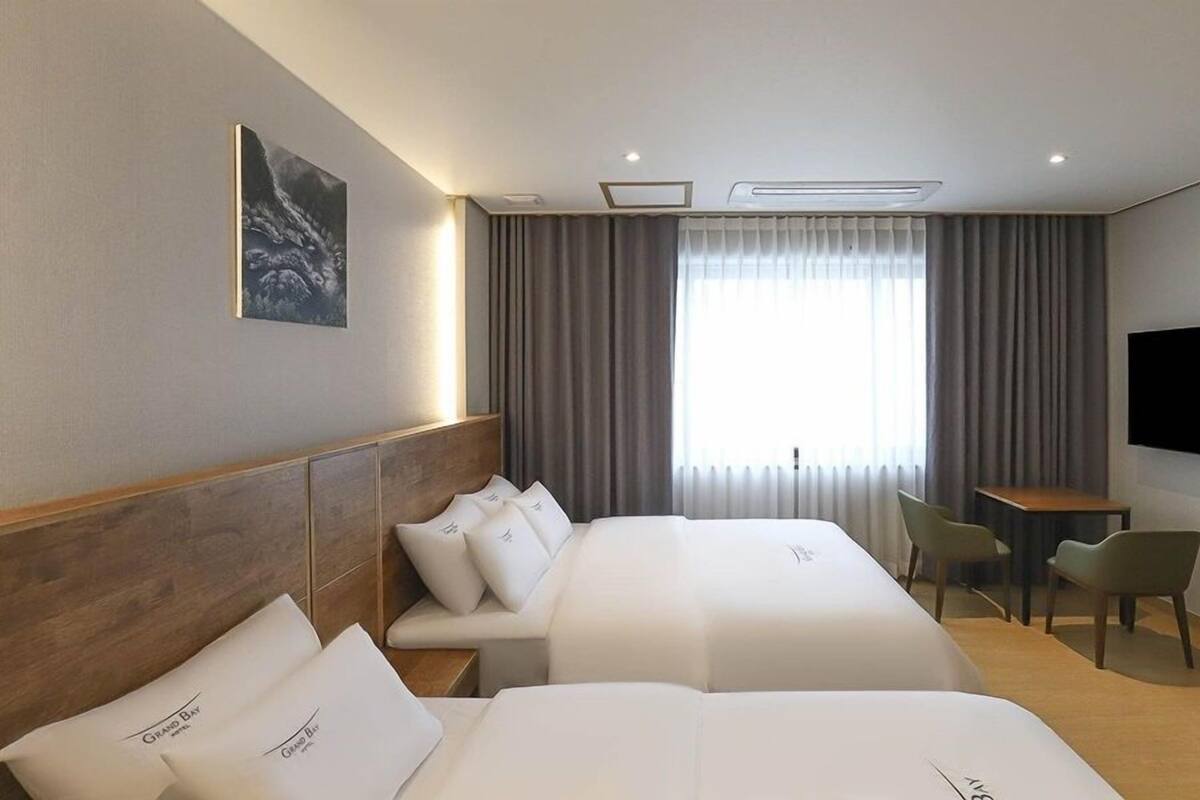 A hotel room with two beds, a desk, a chair, a flat-screen TV, and a large window with curtains.