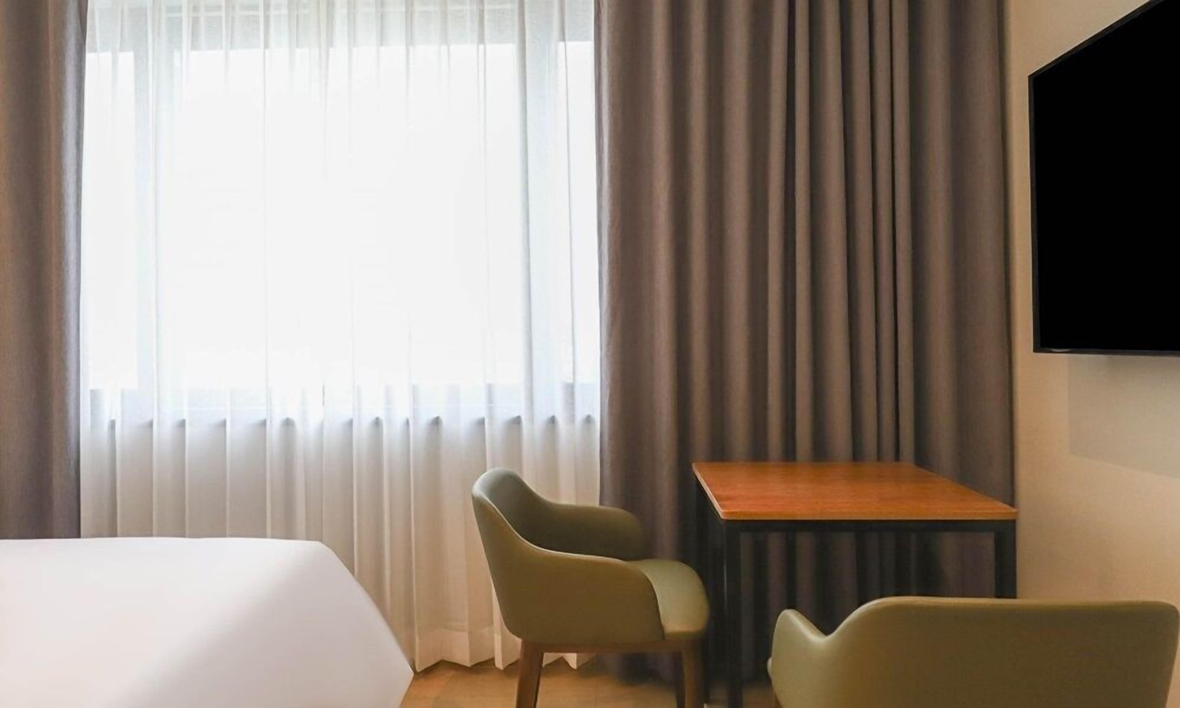 Habitación de hotel con cama, dos sillas, una mesita, televisión y ventana con cortinas.