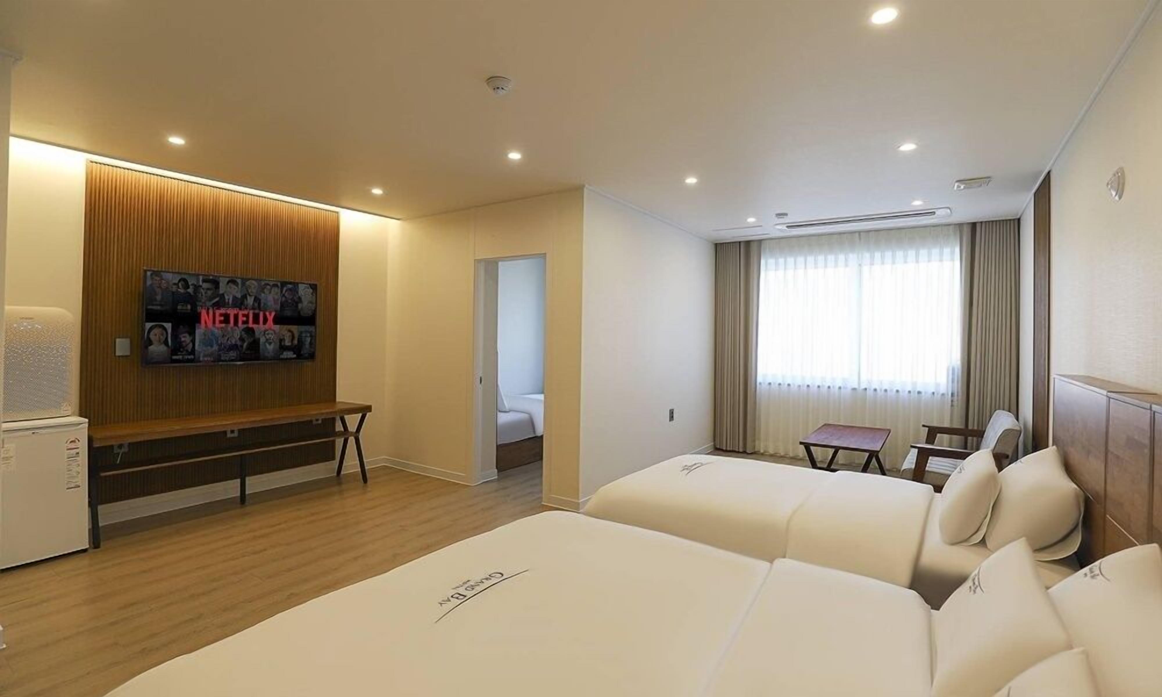 A hotel room with two beds, a TV mounted on the wall, a small table, and a window with curtains.