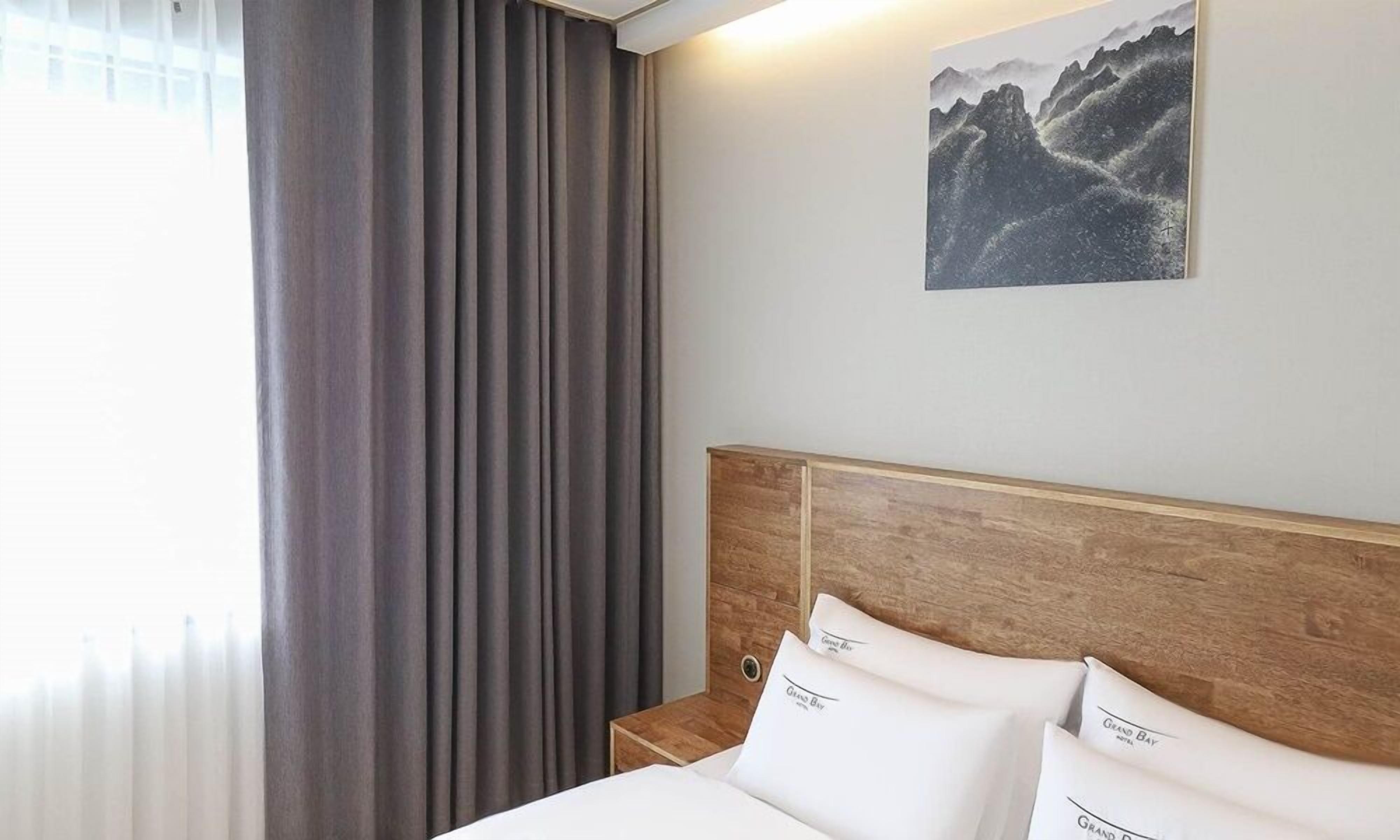 Una habitación de hotel con cabecera de madera, una cama con ropa blanca, una mesita de noche, un cuadro de montañas y una ventana con cortinas.