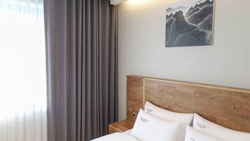 Una habitación de hotel con cabecera de madera, una cama con ropa blanca, una mesita de noche, un cuadro de montañas y una ventana con cortinas.