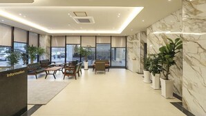 A modern reception area with a marble wall, a reception desk, seating area, and large windows. - Hotel Grand Bay Boryeong (Boryeong)