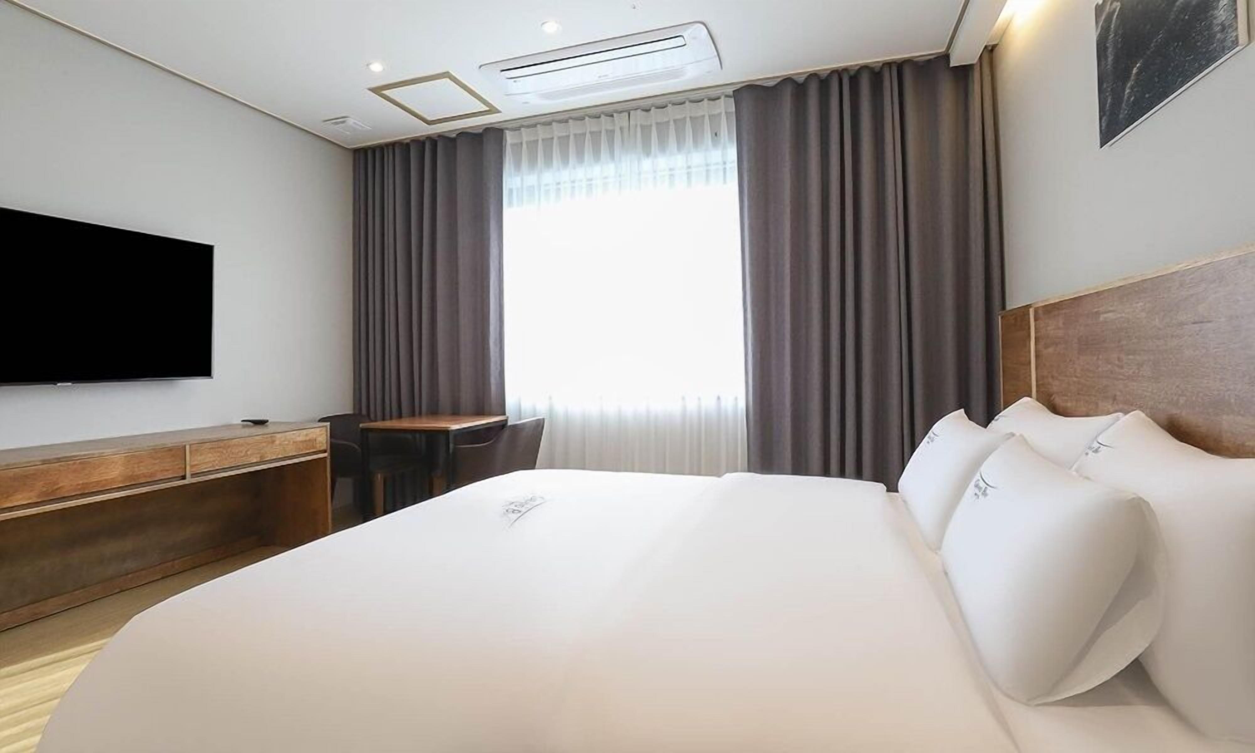 A hotel room with a large bed, a flat-screen TV, a wooden desk, and a window with curtains.