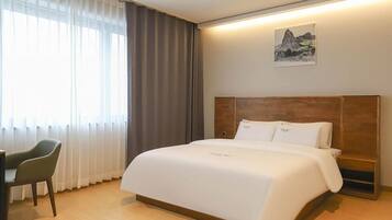 Hotellihuone, jossa on suuri sänky, puinen pääty, työpöytä ja tuoli. Huoneessa on ikkuna, jossa on verhot, ja seinällä on taulu.