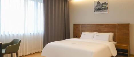 A hotel room with a large bed, a wooden headboard, a desk, and a chair. There is a window with curtains and a painting on the wall.