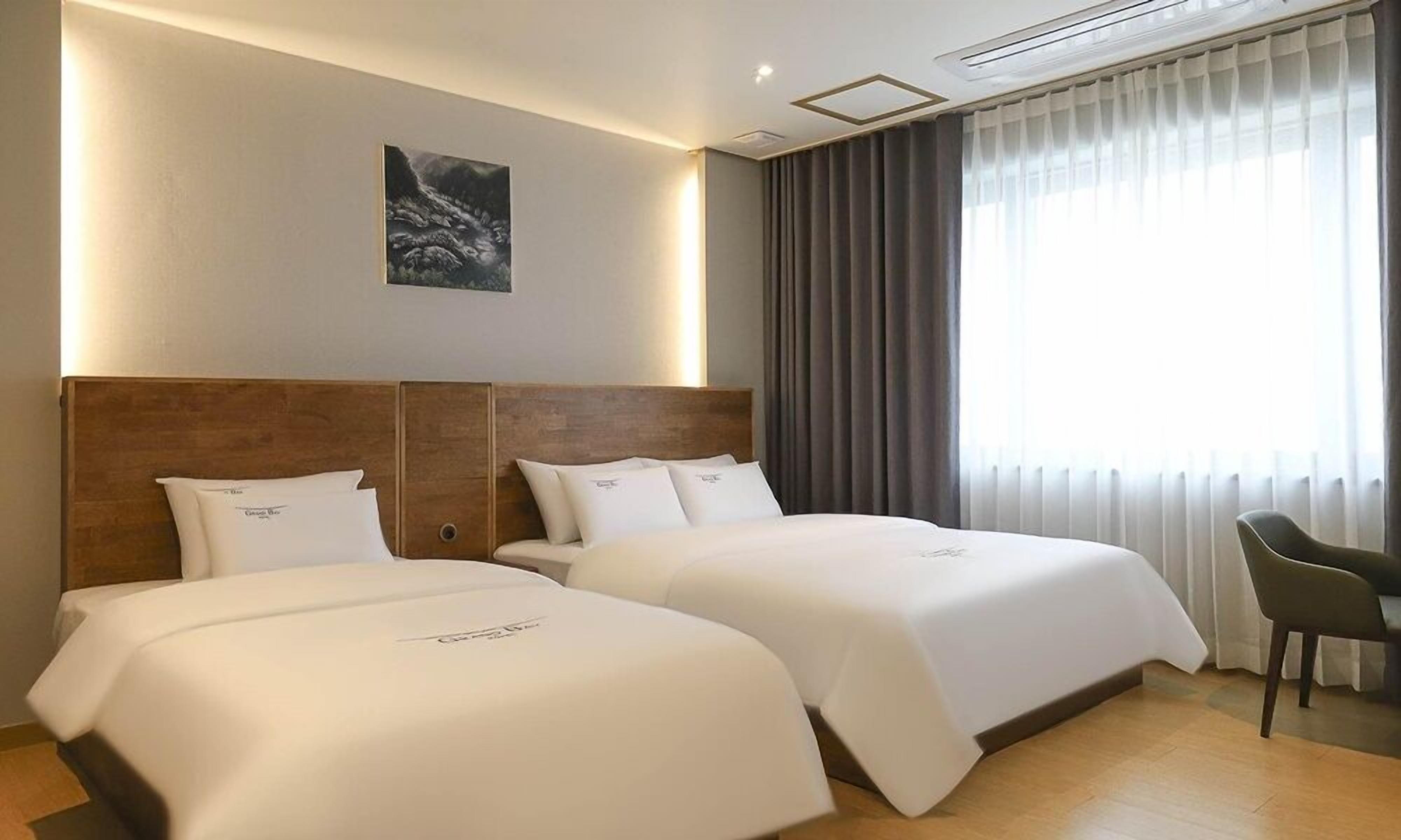A hotel room with two beds, a wooden headboard, a painting on the wall, and a large window with curtains.
