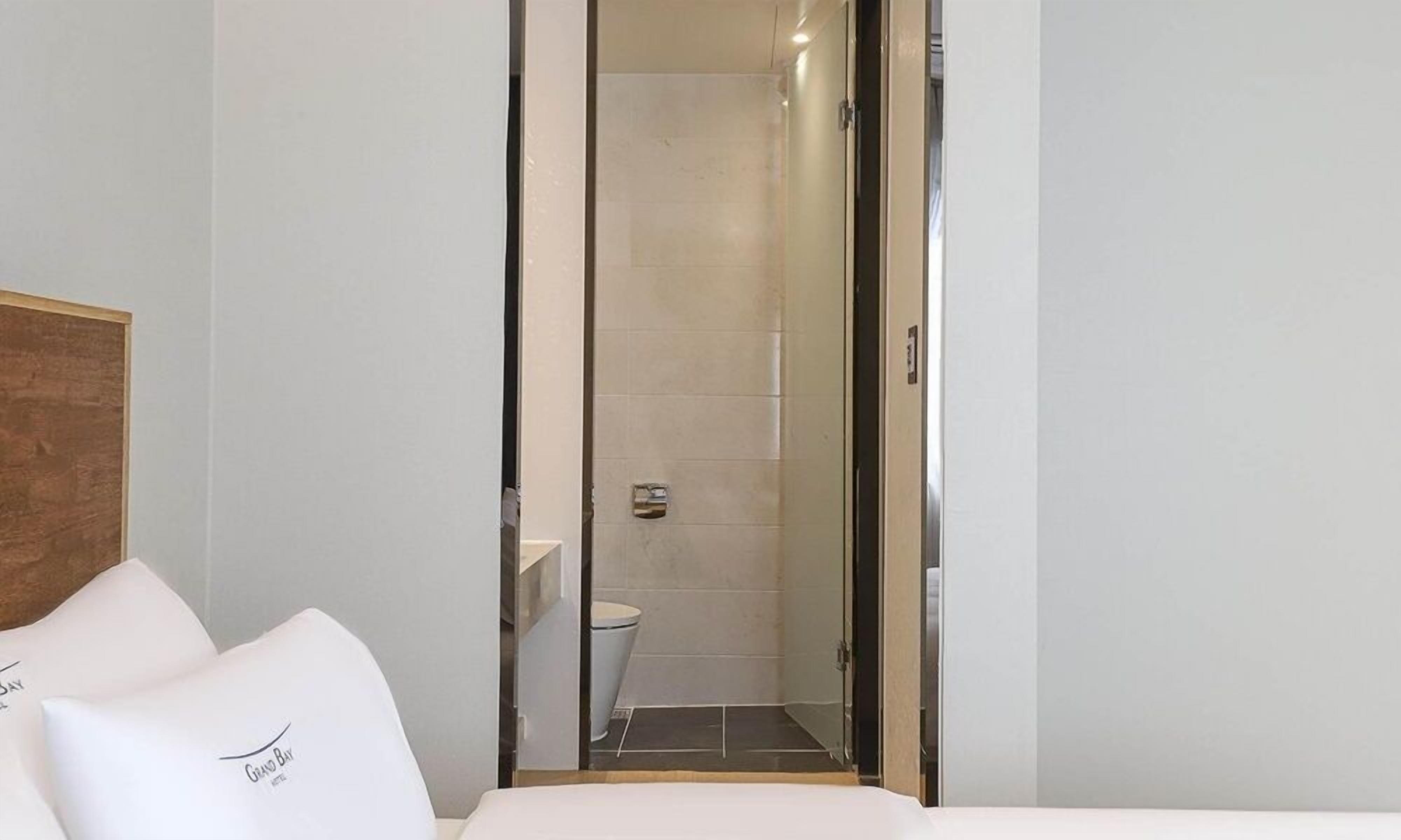 A hotel room with a bed, a wooden headboard, and a glass door leading to a bathroom with a toilet and a shower.