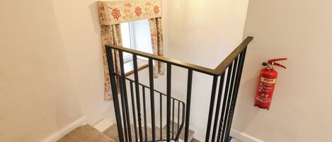 A staircase with a black railing, a window with floral curtains, a wall-mounted light, and a red fire extinguisher.