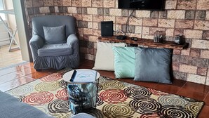A living room with a sofa, a coffee table, a patterned rug, a TV mounted on a brick wall, and a small wooden shelf with decorative items.