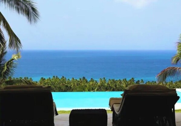 A view of a beach with a clear blue ocean and a pool. There are lounge chairs and palm trees visible.