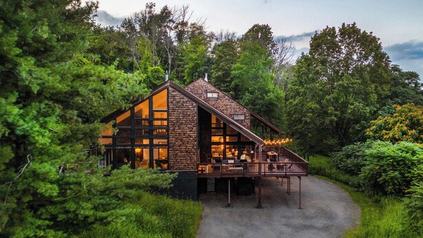 A rustic cabin with a large deck and multiple windows, surrounded by dense forest.
