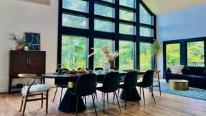 A dining area with a large table, chairs, and a view of greenery through large windows.