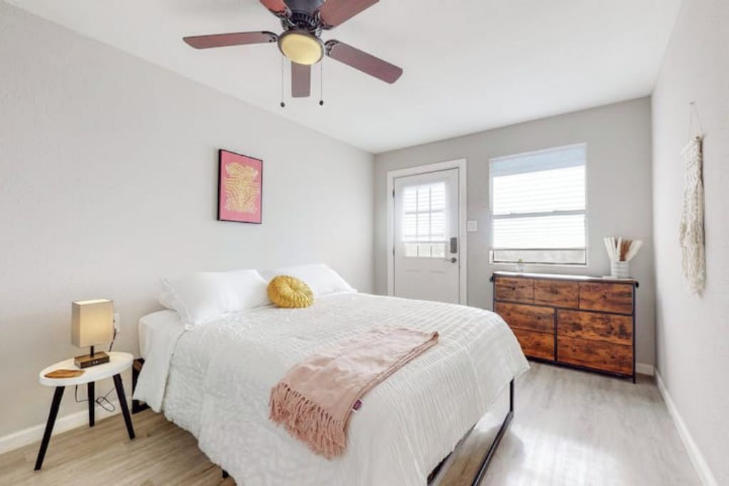 Une chambre à coucher comprenant un lit, un ventilateur de plafond, une table de chevet avec une lampe, une commode et une fenêtre avec des stores.