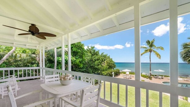 A covered outdoor area with a table and chairs, a ceiling fan, and a view of the ocean and palm trees.