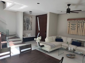 A modern living room with a glass coffee table, a sofa set, a dining table with chairs, and a staircase with glass railings.