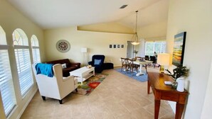 A spacious living room with a sofa, armchair, and dining table. There are windows with blinds, a chandelier, and a painting on the wall. - Villa Gone Golfing (Rotonda West)