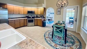 A modern kitchen with wooden cabinets, stainless steel appliances, and a dining area with teal chairs and a patterned rug. - Villa Gone Golfing (Rotonda West)