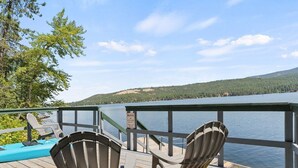 Een houten terras met Adirondack-stoelen met uitzicht op een meer, een waarschuwingsbord en een bebost heuvel.