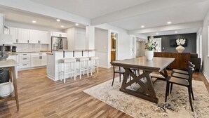 Een ruime keuken met witte kastjes, een centraal eiland met barstoelen, een eethoek met een houten tafel en stoelen, en een zithoek met een bank en decoratieve vaas.