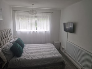 A bedroom with a bed, a television, a radiator, and a window with sheer curtains.