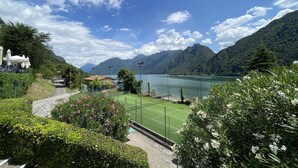 A tennis court with a mountain backdrop, surrounded by lush greenery and a clear sky. - Directly on the lake with terrace (Tre Capitelli)