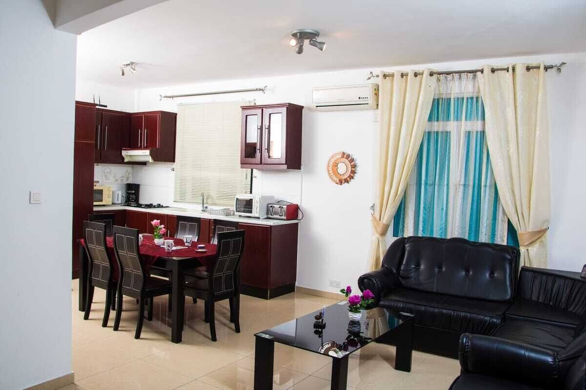 A compact living space with a dining area, a kitchenette, a black leather sofa, and a window with curtains.