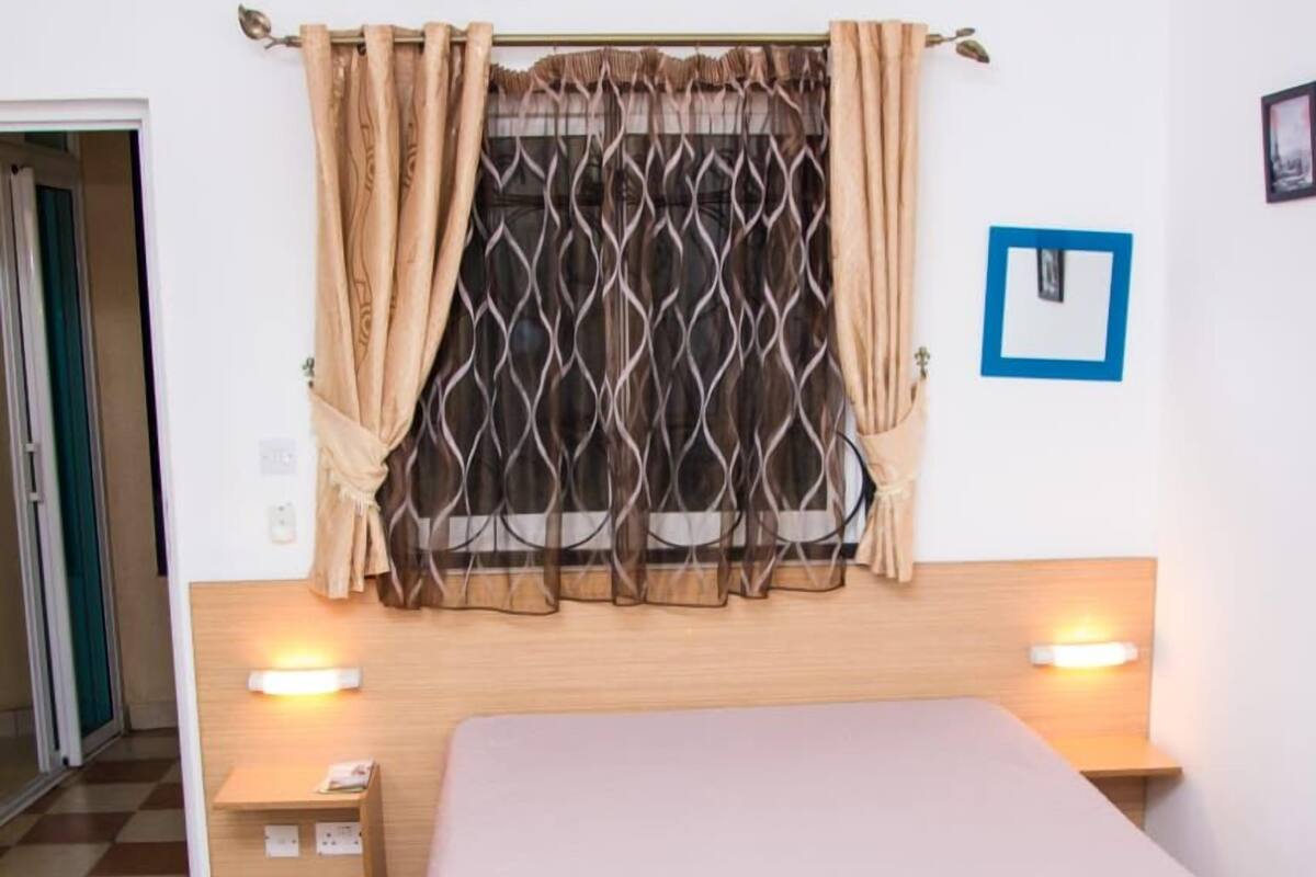 A bedroom with a bed, bedside lamps, a chandelier, a window with curtains, and a framed picture on the wall.