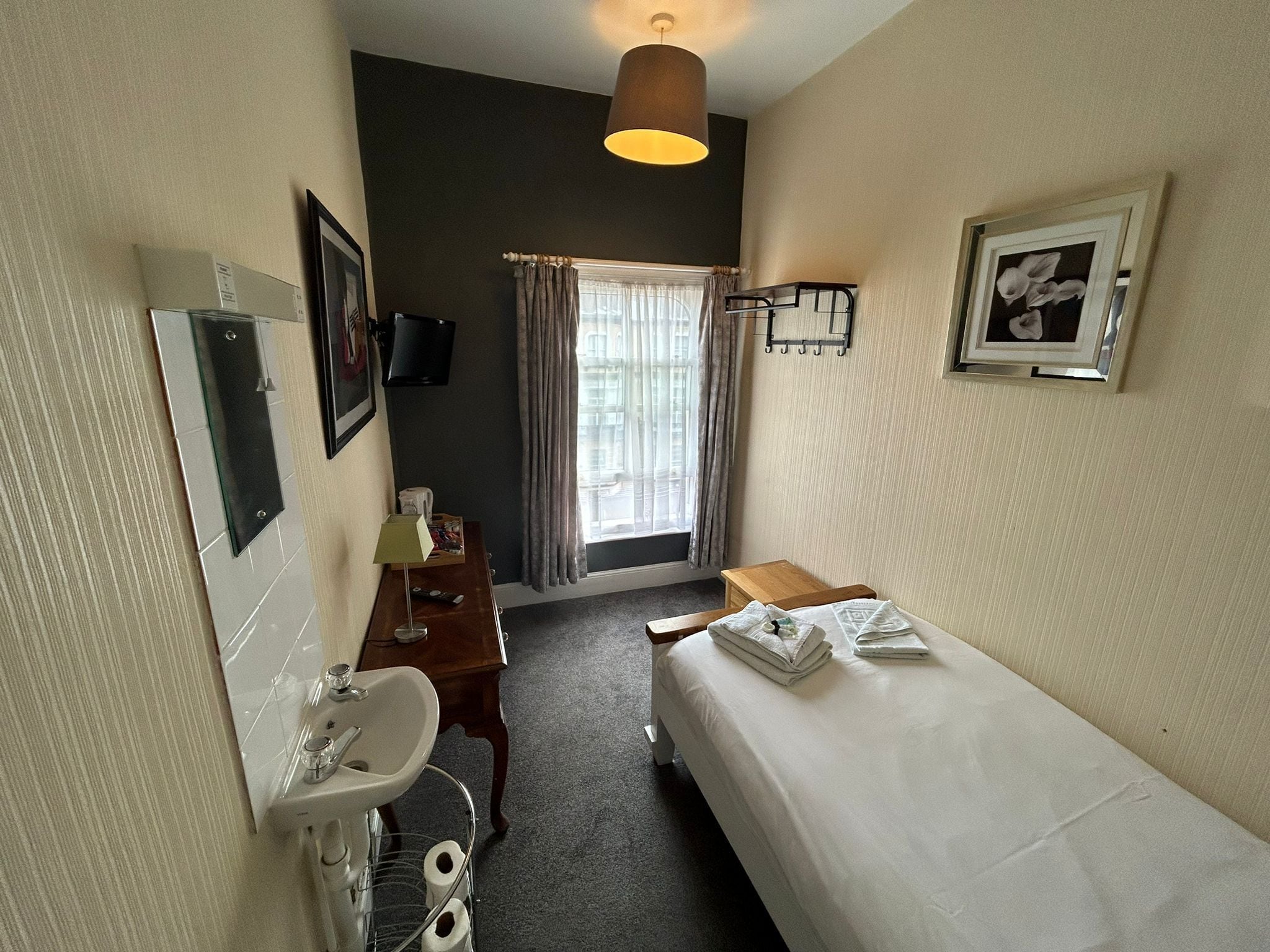 Una habitación de hotel pequeña y bien iluminada, con una cama individual, una mesita de noche, un escritorio con silla, un lavamanos y un espejo.