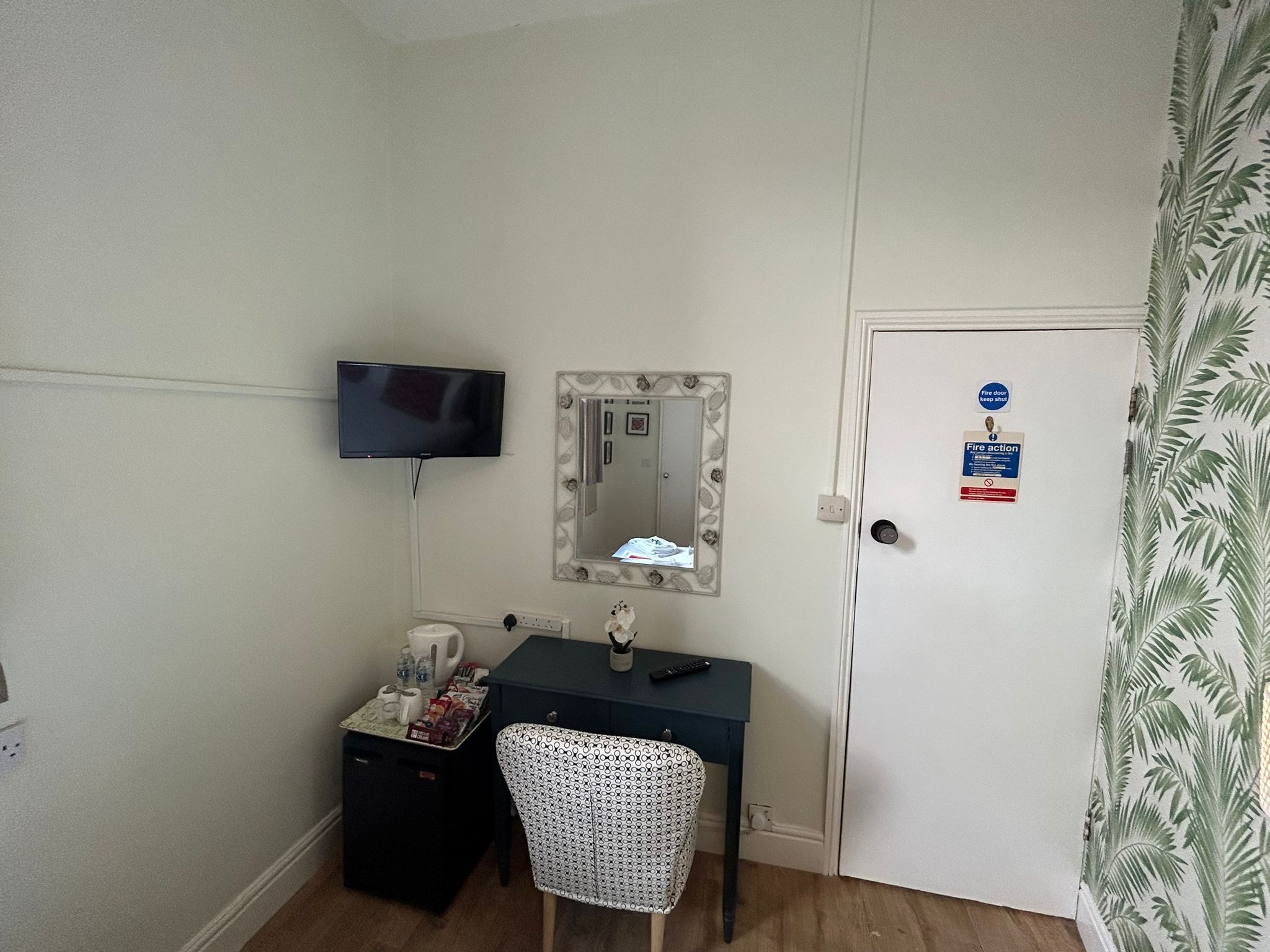 Ein kleines Zimmer mit Schreibtisch, Stuhl, Spiegel, Fernseher und einer Tür mit einem Brandschutzschild.