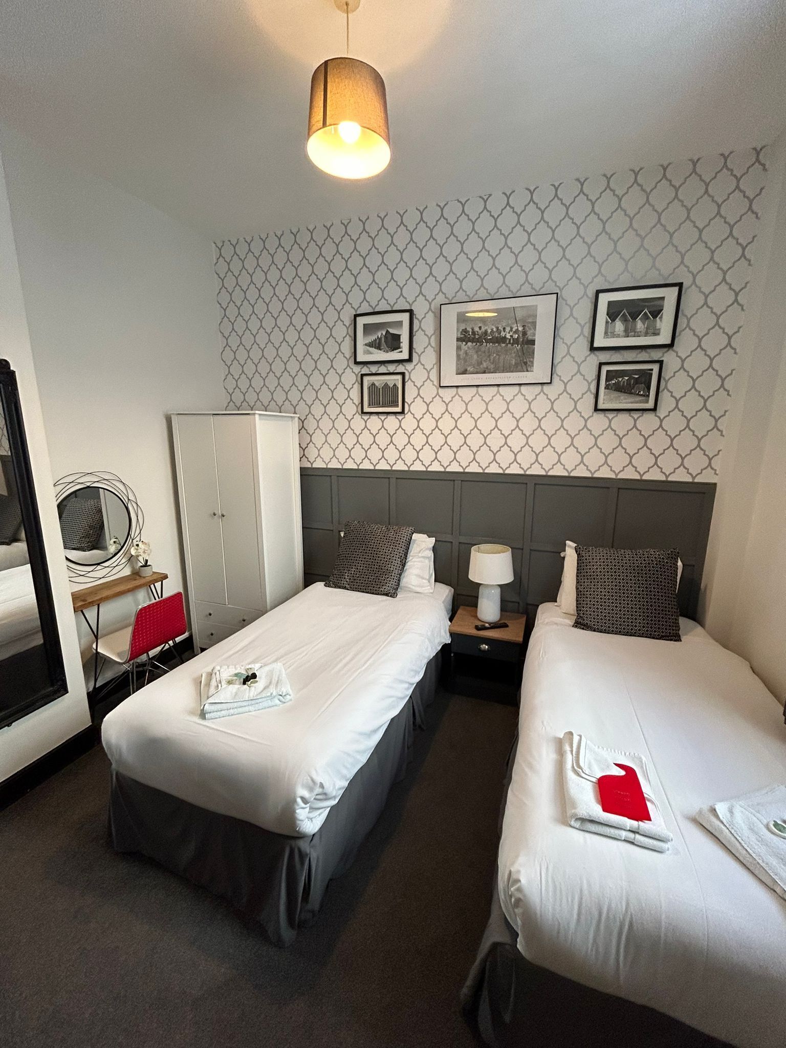 A hotel room with two single beds, a wardrobe, a desk with a chair, a mirror, and a wall with framed pictures.