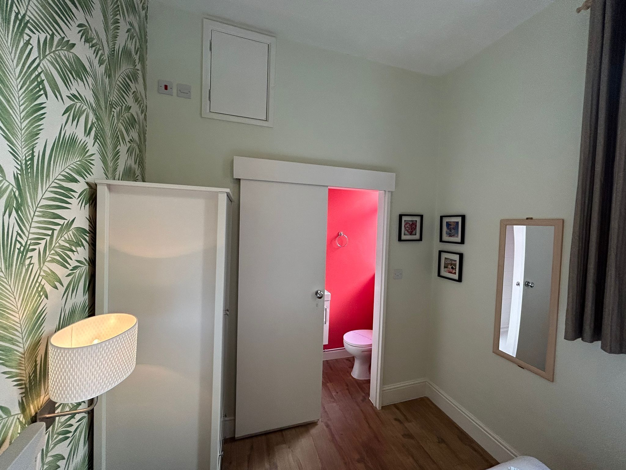 Une pièce avec une porte blanche donnant sur une salle de bain dont un mur est rouge et où se trouve des toilettes blanches. Un miroir est accroché au mur et une lampe se trouve sur le côté gauche.