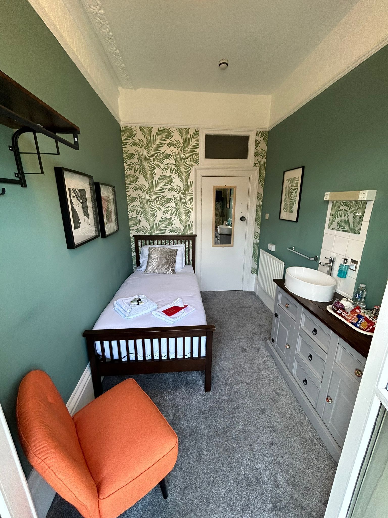 Habitación compacta con cama, una silla, baño con lavamanos y espejo, y una puerta que da a otra habitación.