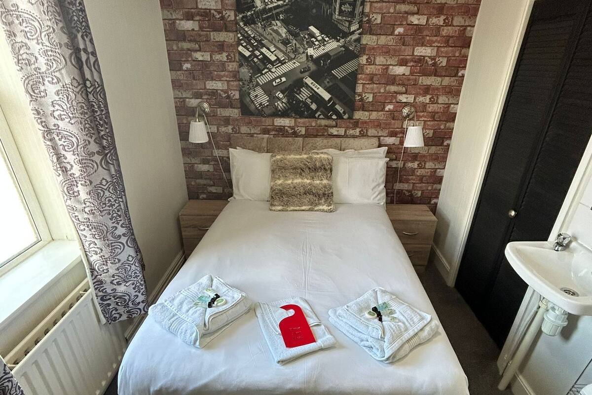 Un lit bien fait, avec une étiquette rouge, un mur de briques orné d’une image représentant un paysage urbain, un lustre et une salle de bain avec un lavabo et des toilettes.