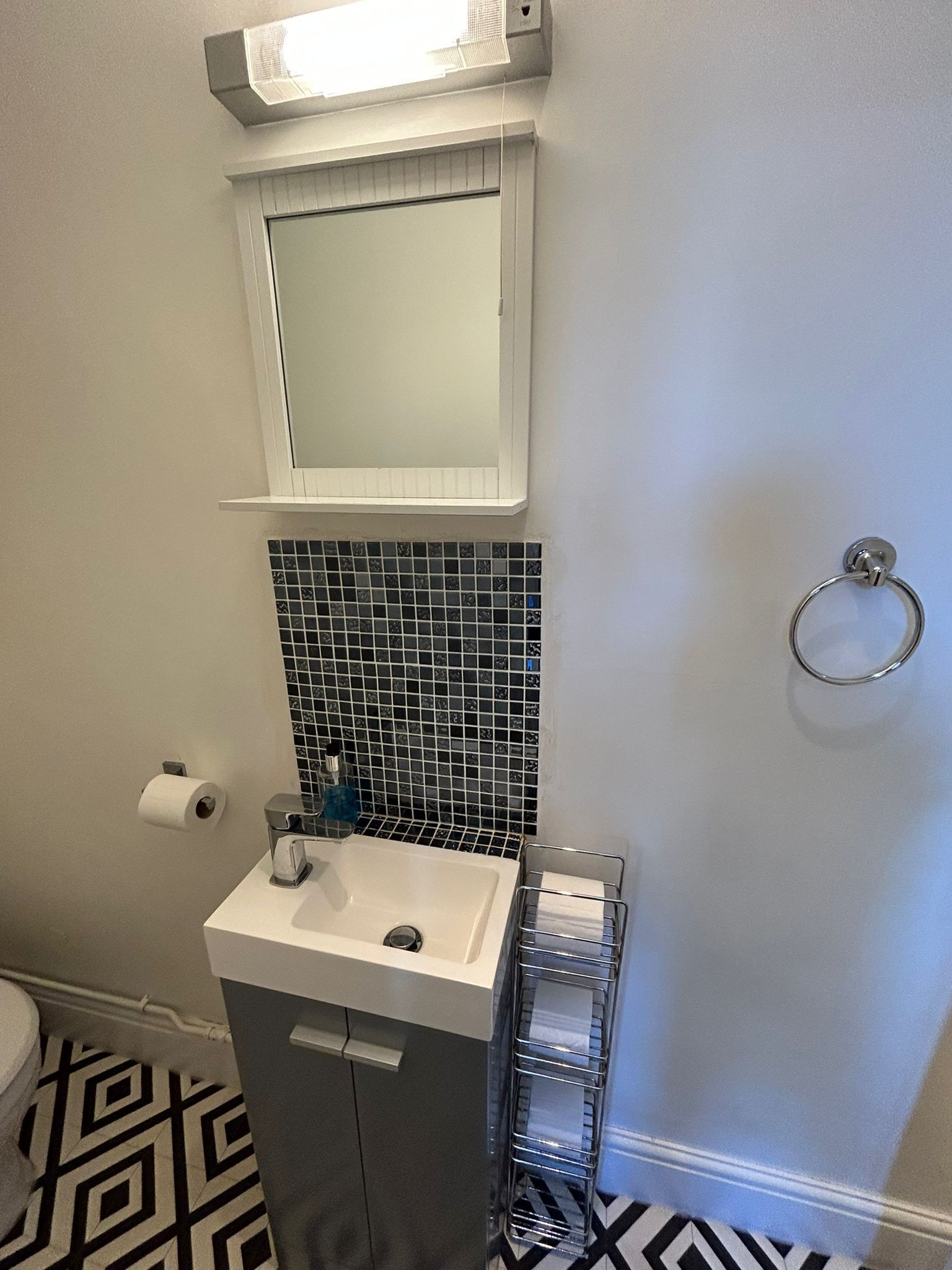 Et badeværelse med en hvid vask, et spejl over vasken, et toilet og et gulv med et mønster.