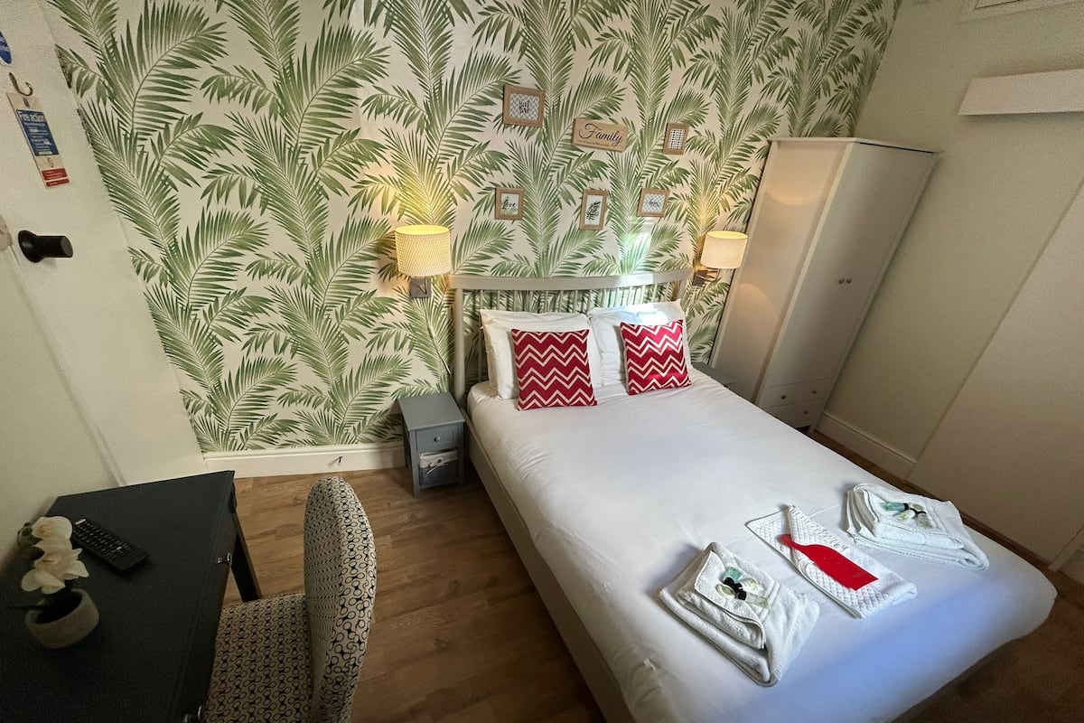 Une chambre d’hôtel comprenant un lit, des lampes de chevet, un bureau avec une chaise et un mur recouvert d’un papier peint à motifs de feuilles de palmier.