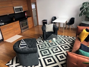 A modern kitchen with wooden cabinets, a microwave, and a refrigerator. There is a black sofa with a patterned cushion, a glass coffee table, and a black and white rug. A small table with a white tablecloth and black chairs is visible in the background.