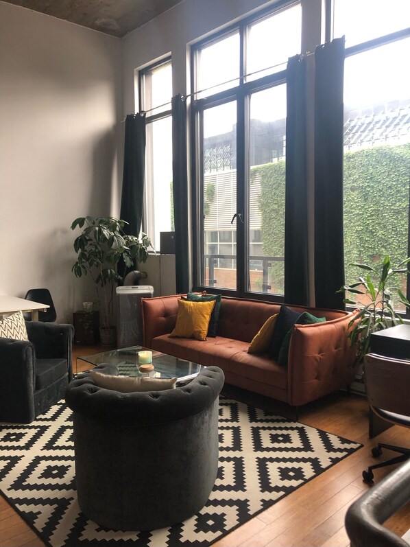 A living room with a sofa, ottoman, and a glass coffee table. There are large windows with curtains, a potted plant, and a patterned rug.