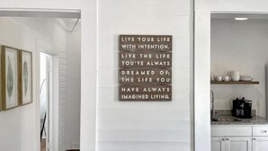 A hallway with a motivational quote on the wall, a kitchen area with white cabinets and a coffee maker, and framed artwork on the wall. - Best neighborhood in SA! Near it all, food & fun!
 (San Antonio)