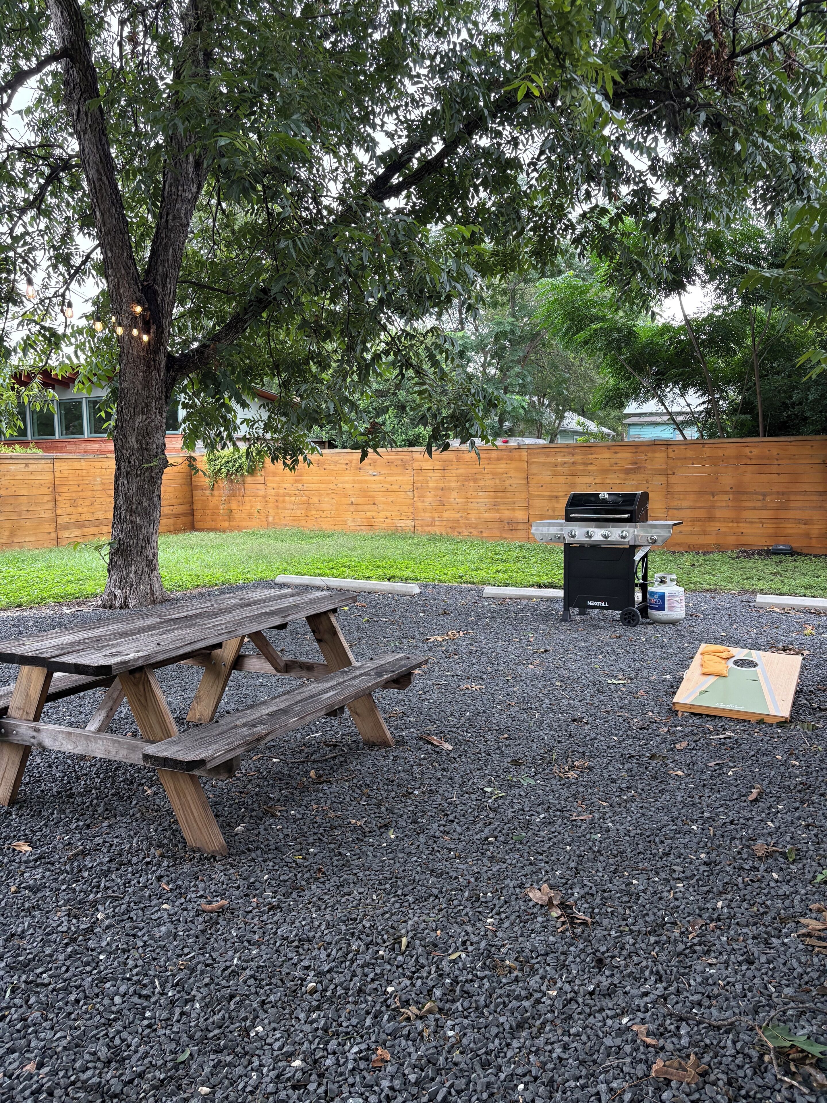 A picnic area with a wooden table and a barbecue grill, surrounded by a wooden fence and trees.