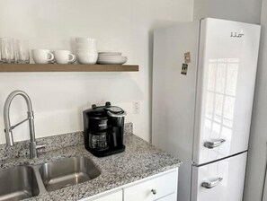 Uma cozinha com pia dupla, cafeteira, geladeira branca e prateleira com xícaras e pratos.