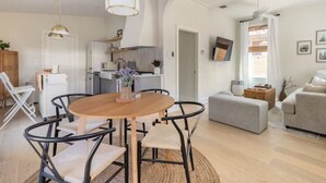 Ein modernes Wohnzimmer mit einem runden Holztisch, schwarzen Metallstühlen, einem grauen Sofa und einem weißen Sessel. Es gibt eine Küchenzeile mit weißen Schränken und einer Mikrowelle.