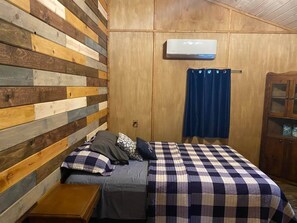 Una camera da letto con parete a doghe di legno, un letto con copriletto a scacchi, un comodino, un armadietto in legno e un condizionatore d'aria.