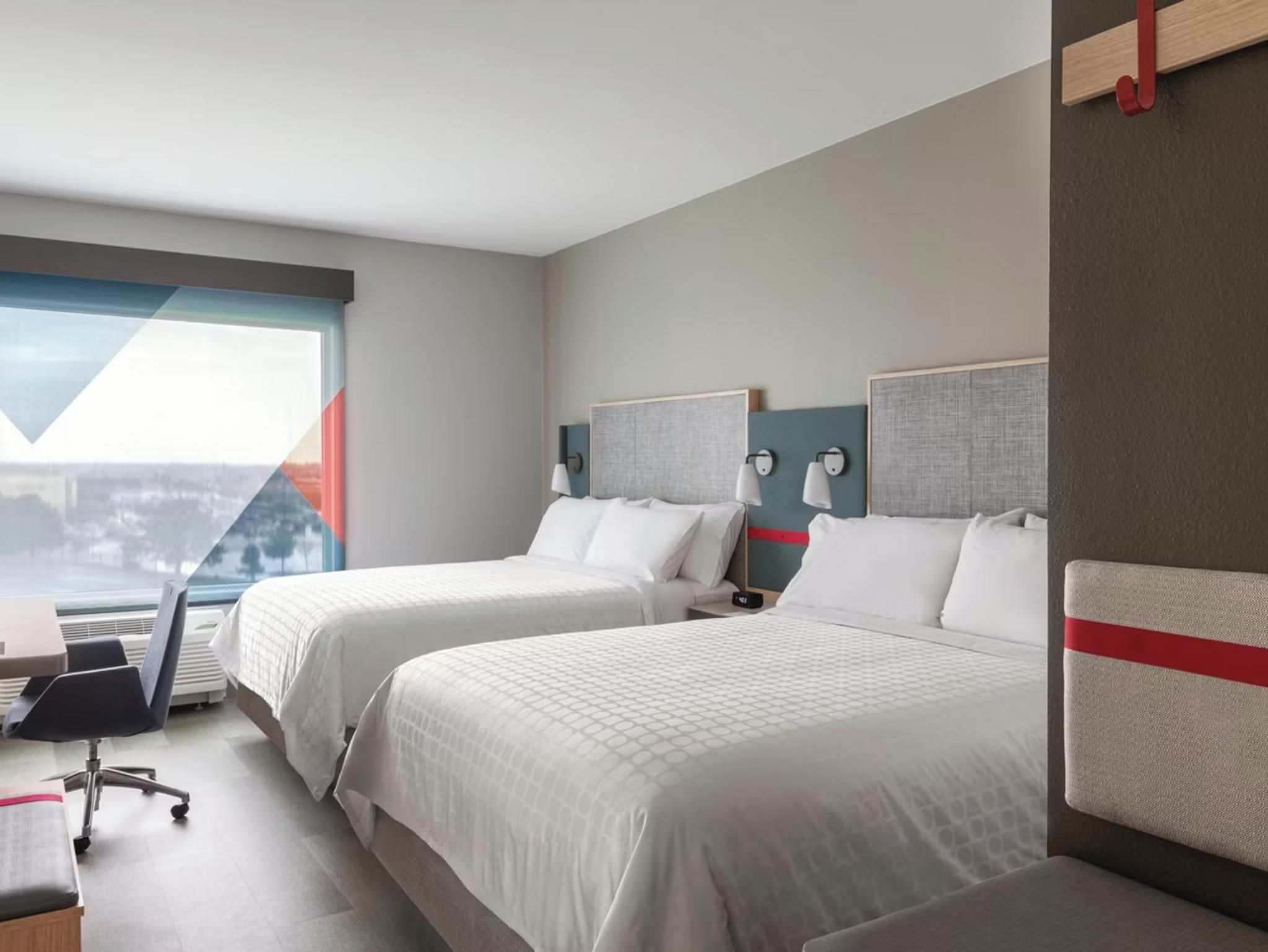 A hotel room with two beds, a desk, a chair, and a view of a cityscape.