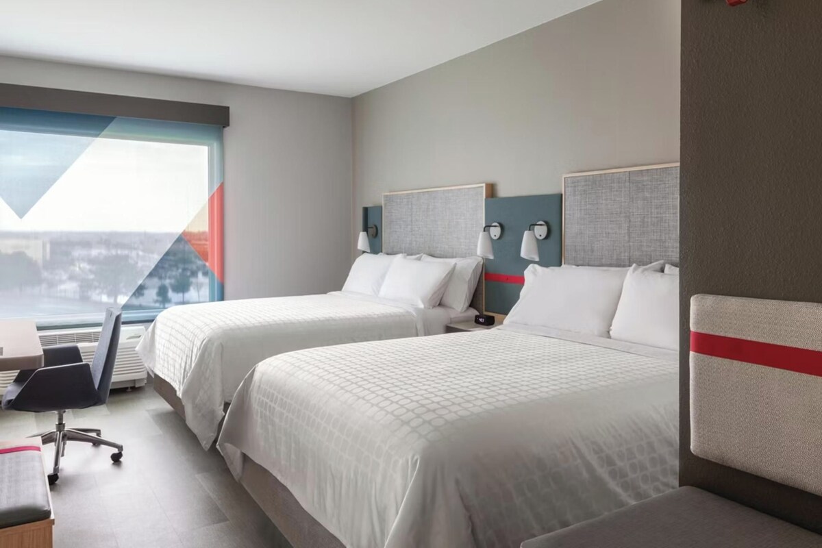A hotel room with two beds, a desk, a chair, and a view of a cityscape.