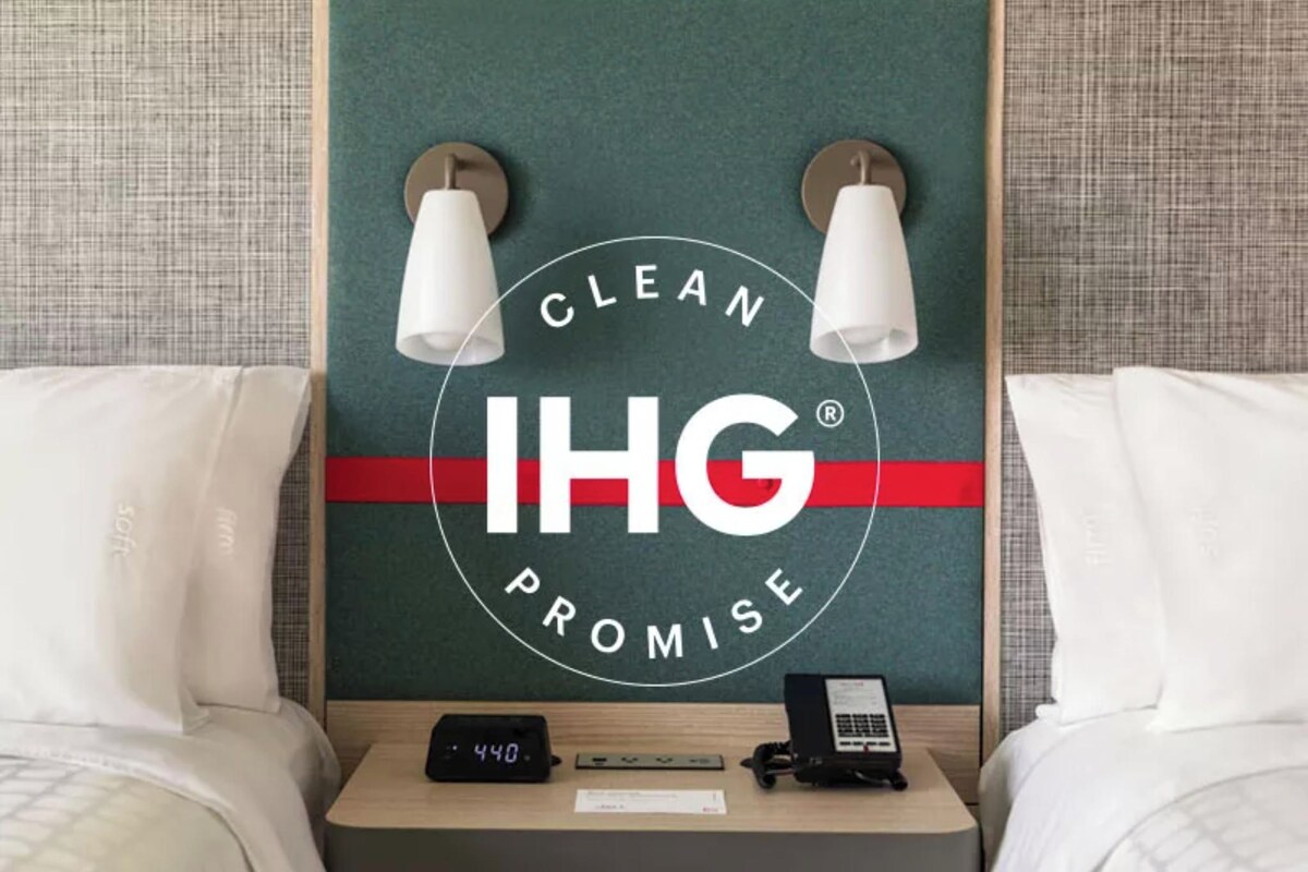 A hotel room with a headboard displaying the logo 'CLEAN IHG PROMISE' and two wall-mounted lamps.