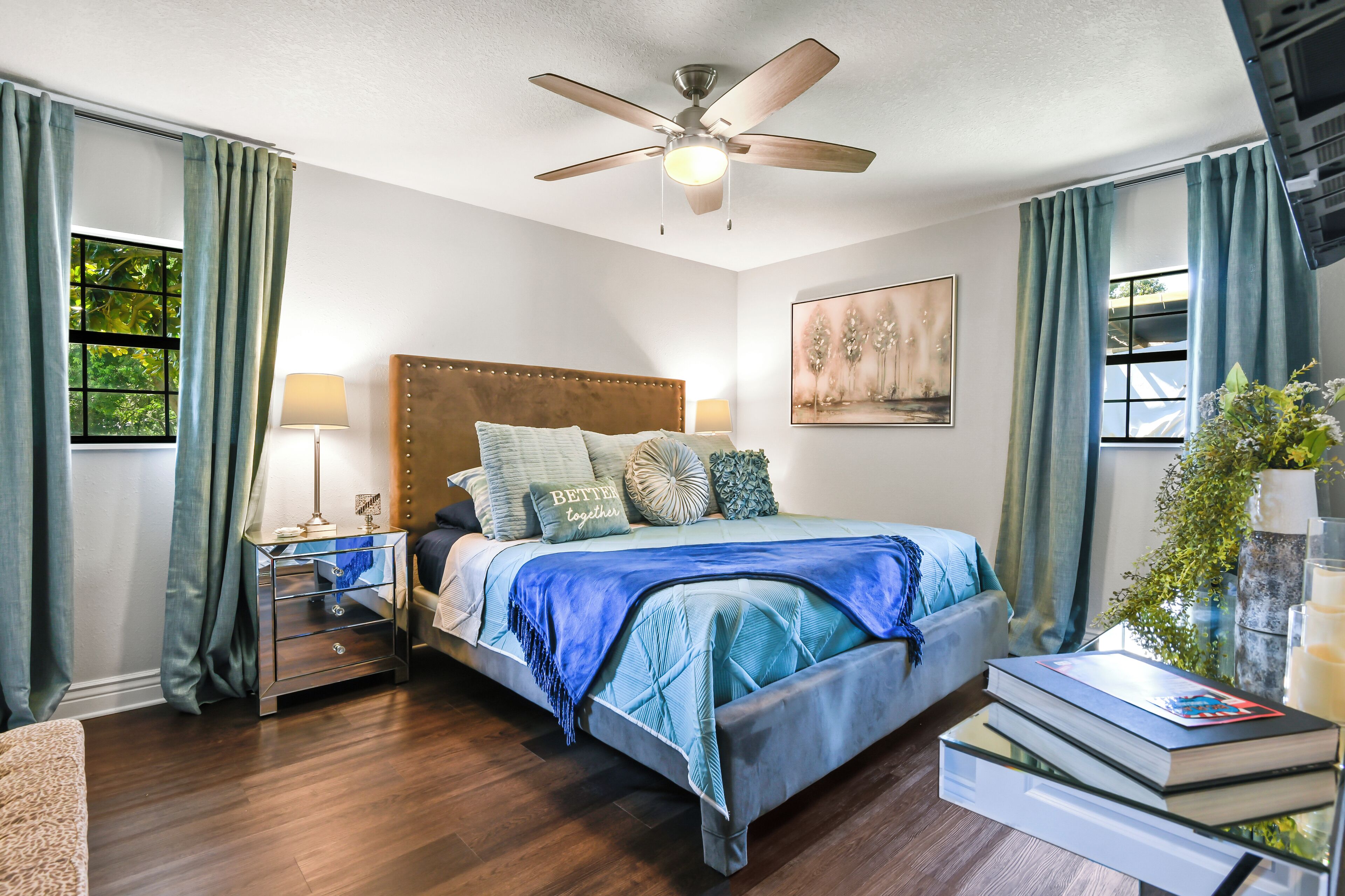 Une chambre à coucher comprenant un lit, des tables de chevet, un ventilateur de plafond, une fenêtre avec des rideaux, une table de chevet, une lampe, une plante sur la table de chevet et un tableau mural.