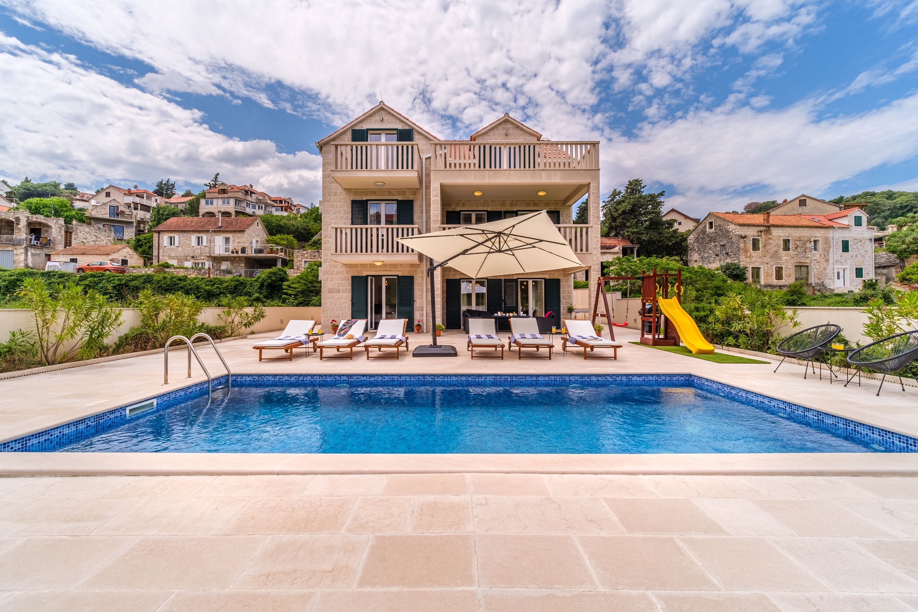 A swimming pool with lounge chairs and a slide, surrounded by a building with balconies and a view of other houses.