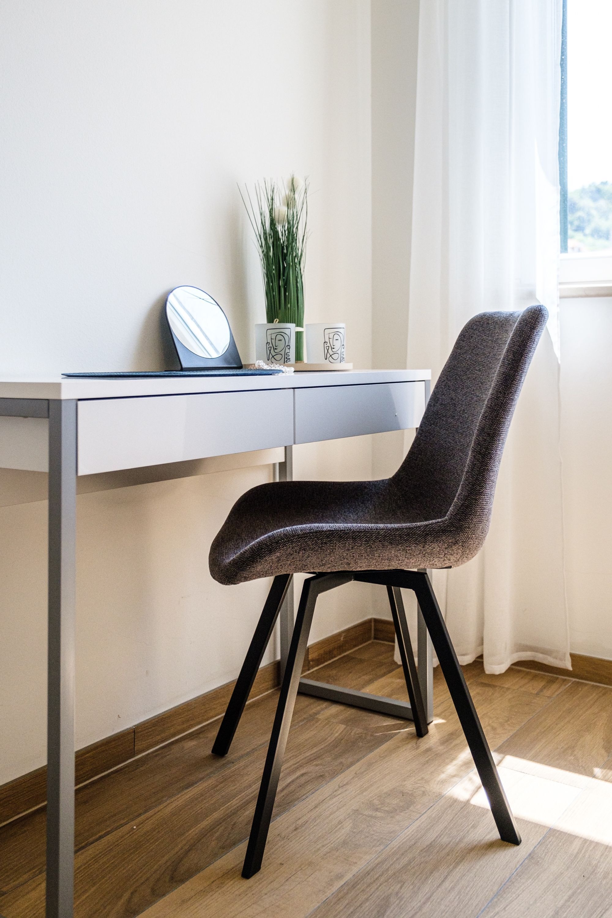 Een moderne bureaustoel met een donkere stoffen zitting en rugleuning, geplaatst voor een wit bureau met een laptop, een ronde spiegel en een kleine potplant.