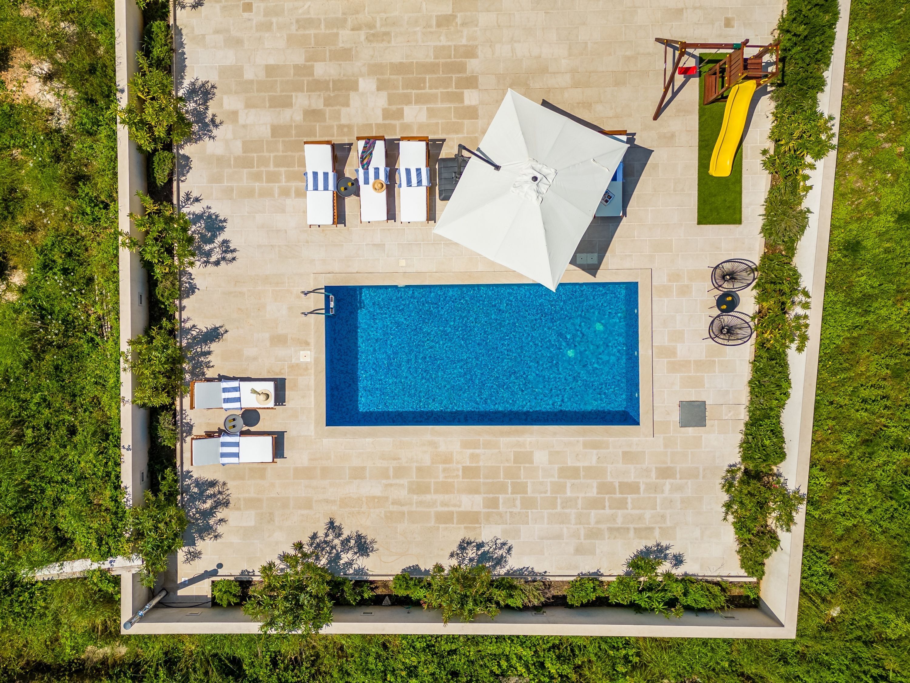 An aerial view of a swimming pool area with a rectangular pool, lounge chairs, umbrellas, and a children's slide.