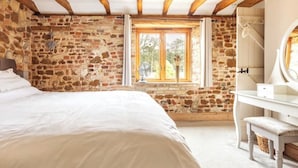 Un dormitori amb paret de pedra, un llit gran, sostre de fusta, una finestra amb cortines i un envellidor blanc amb mirall.